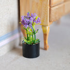 1:12 scale miniature potted flowering plant with purple blooms in a black round pot, shown on the floor by a dresser