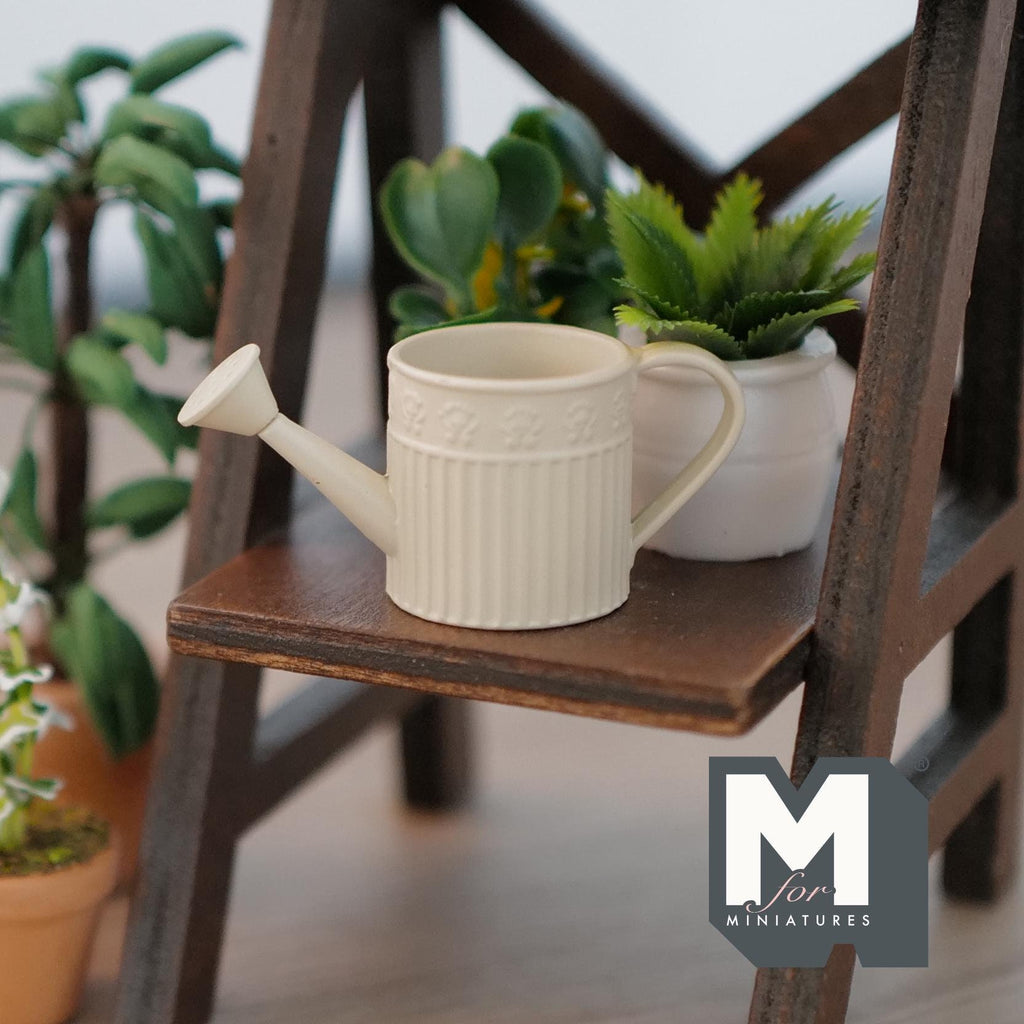 1:12 scale miniature cream metal round watering can with ribbed body, shown on a dark wood plant shelf with potted plants