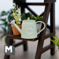 1:12 scale miniature metal slanted watering can in light green with a ribbed body, shown on a dark wood plant stand