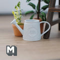 1:12 scale miniature metal slanted watering can in light blue with embossed pattern, shown beside potted plants