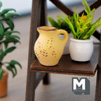1:12 scale miniature clay watering jug in pale yellow with handle and spout, on a wood plant stand beside a potted plant
