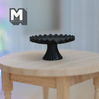 1:12 scale miniature metal pedestal cake stand in black with a scalloped rim, shown on a round wood table