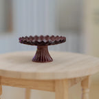 1:12 scale miniature metal pedestal cake stand in brown with a scalloped rim and fluted base, shown on a round wood table