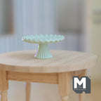 1:12 scale miniature metal pedestal cake stand in light green with a fluted rim, shown on a round wood table