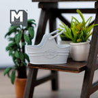 1:12 scale miniature metal flower basket in light blue with arched handle, shown on a wood stand beside potted plants