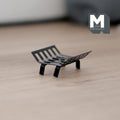 1:12 scale miniature metal fire grate in black with curved slatted bars and four legs, shown on a light wood floor