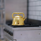 1:12 scale miniature gold metal kettle with lid and twisted handle, shown on a kitchen stove top