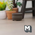 1:12 scale miniature short round clay tree pot in dark gray, shown on a wood floor beside potted plants
