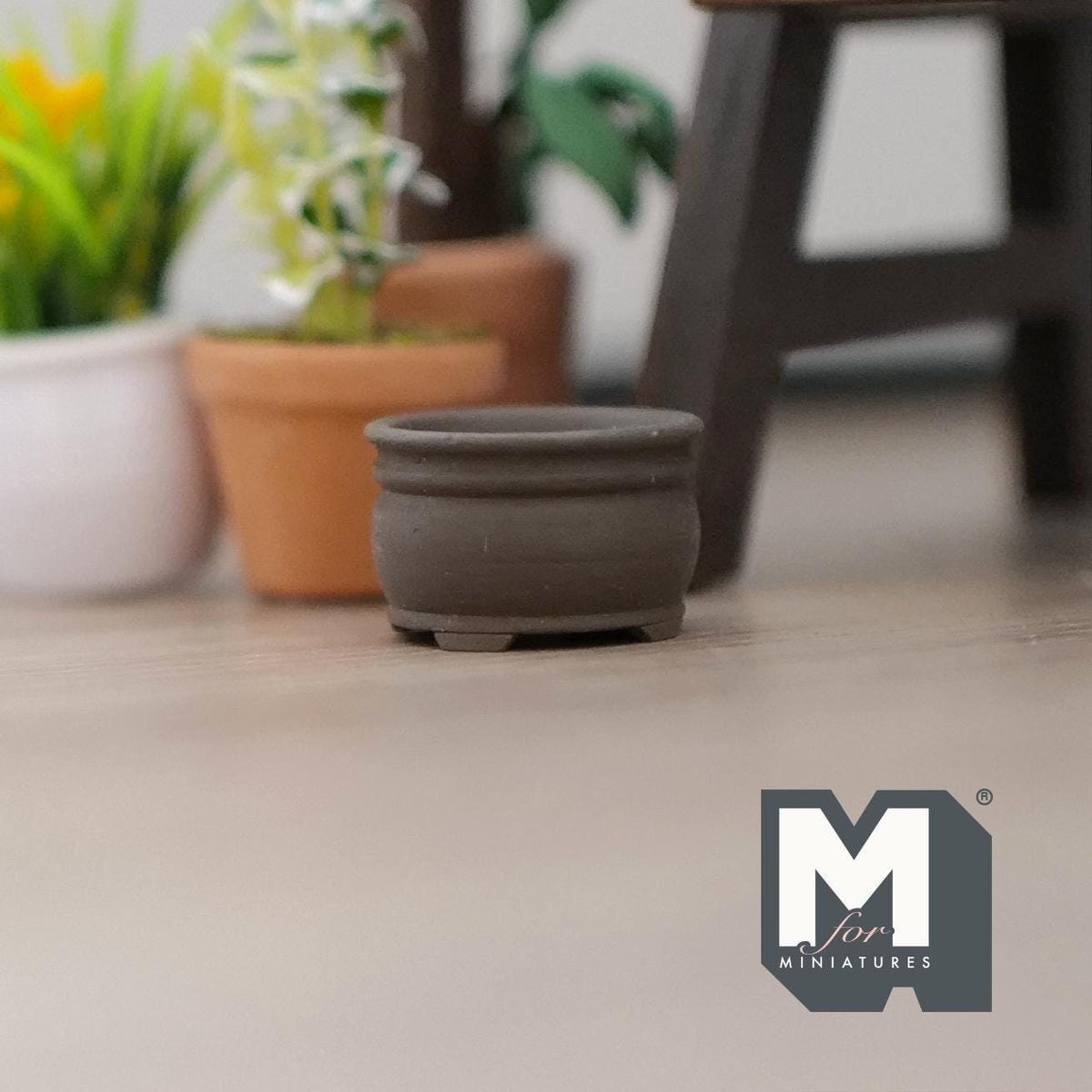1:12 scale miniature short round clay tree pot in dark gray, shown on a wood floor beside potted plants