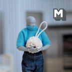1:12 scale miniature shower loofah in beige with a white hanging cord, held by an artist figure