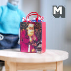 1:12 scale miniature Christmas gift bag with red rope handles and a Santa print, shown on a round wood table