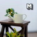 1:12 scale miniature light green metal round watering can with ribbed body, shown on a dark wood plant stand