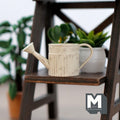 1:12 scale miniature metal round watering can in beige with rustic label and long spout, on a dark shelf by plants