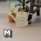 1:12 scale miniature rustic metal watering can in beige with slanted ribbed body and label, beside potted plants