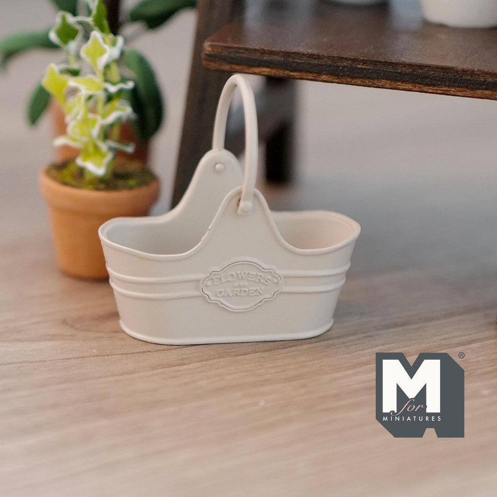 1:12 scale miniature metal flower basket in beige with arched handle and embossed label, on a wood floor by a potted plant