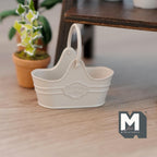 1:12 scale miniature metal flower basket in beige with arched handle and embossed label, on a wood floor by a potted plant