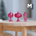 1:12 scale miniature footed candy dishes with lids in clear magenta, set of 3 shapes, on a round wood table