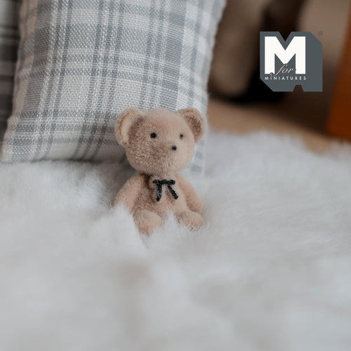 1:12 scale miniature beige bear cub with bow tie and flocked skin, sitting on a white fur rug beside a plaid pillow