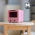 1:12 scale miniature metal toaster oven in pink with clear door and two front knobs, shown on a round wood table