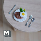 1:12 scale miniature steak and fried egg on a plate with silver knife and fork, shown on a round wood table