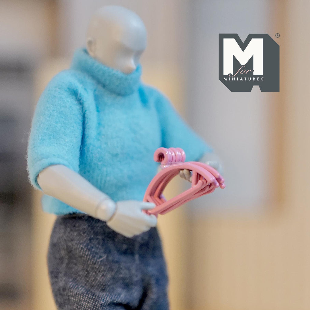 1:12 scale miniature metal clothes hangers in pink, set of 6, shown held by a poseable figure