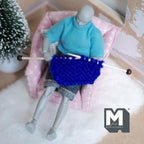 1:12 scale miniature knitting needles with blue knitted piece, shown held by a jointed figure seated in a pink armchair