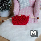 1:12 scale miniature knitting needles with a red knitted piece, shown on a white fluffy rug beside a pink armchair