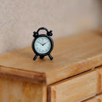 1:12 scale miniature metal twin bell alarm clock in matte black with white dial, shown on a wood dresser