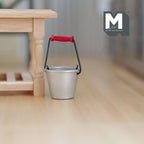 1:12 scale miniature metal bucket in silver with wire handle and red grip, shown on a wood floor by a table