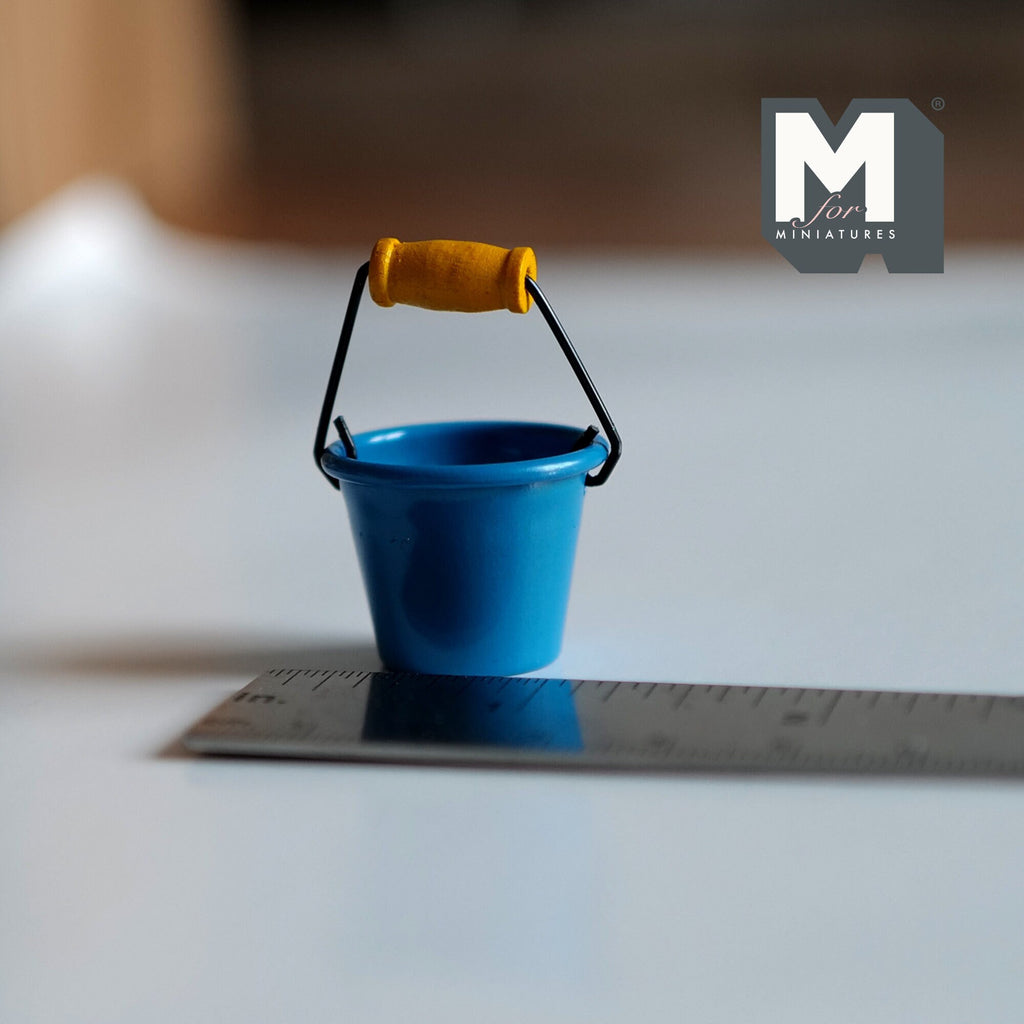 1:12 scale miniature metal bucket in blue with wire handle and wood grip, shown on a ruler for scale