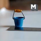 1:12 scale miniature metal bucket in blue with wire handle and wood grip, shown on a ruler for scale