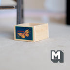 1:12 scale miniature wood fruit crate with butterfly decal label, natural finish, shown empty on a pale surface