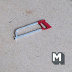 1:12 scale miniature metal hacksaw with red handle and silver frame, shown lying on a brown craft board