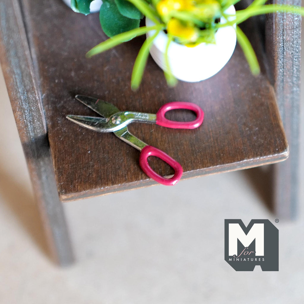 1:12 scale miniature metal tin snips with red handles, non working, shown on a brown wood shelf by a plant