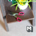 1:12 scale miniature metal tin snips with red handles, non working, shown on a brown wood shelf by a plant
