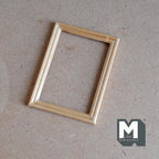 1:12 scale miniature wood picture frame in natural finish, rectangular and open, shown lying on a brown craft board