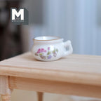 1:12 scale miniature porcelain chamber pot in white with a pink floral design and side handle, shown on a wood table
