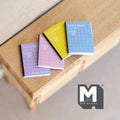 1:12 scale miniature notebooks with pastel grid covers, set of 4, purple, pink, yellow and blue, on a wood table