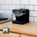 1:12 scale miniature black espresso machine with cup, silver trim, shown on a wood kitchen counter by white tile