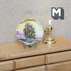 1:12 scale miniature ceramic decorative plate with sailing ship and metal stand, shown on a wood dresser beside a lamp