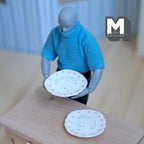 1:12 scale miniature resin large round plates with tiny pink and blue floral pattern, set of 2, held by a jointed figure