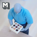 1:12 scale miniature resin box of chocolates with 9 assorted pieces in a white tray, non edible, held by a jointed figure