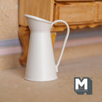 1:12 scale miniature metal pitcher vase in white with a tall handle and spout, shown on the floor beside a wood dresser