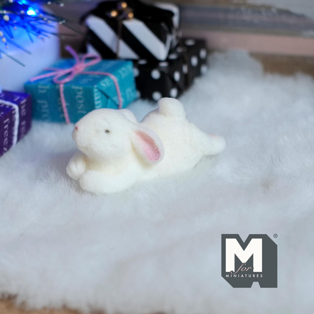 1:12 scale miniature lying bunny figurine in white flocked finish with pink ears, on a white rug by wrapped gifts