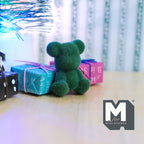 1:12 scale miniature flocked bear cub in dark green, sitting, shown beside wrapped gift boxes on a wood floor