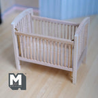 1:12 scale miniature drop side baby crib with pad in bare wood with slatted sides, shown on a wood floor