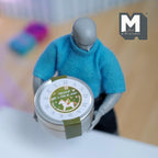 1:12 scale miniature round Christmas tin with lid in silver and green with a label print, shown held by an artist figure