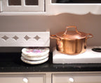 1:12 scale miniature copper plated cooking pot with lid and two handles, shown on a white kitchen stove beside floral plates