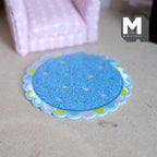 1:12 scale miniature round scalloped shag nursery rug in blue with pastel dots and a pastel edge, shown by a pink armchair