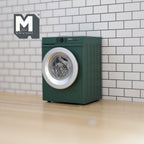 1:12 scale miniature front load washing machine with rotating drum in dark green, shown against a white tile wall